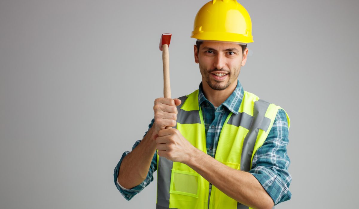 smiling young male engineer wearing safety helmet and uniform holding hammer looking at camera isolated on white background with copy space